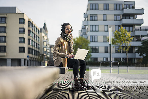 Frau sitzt auf Bank mit Laptop und Kaffee  um mit schnurlosen Kopfhörern Musik zu hören