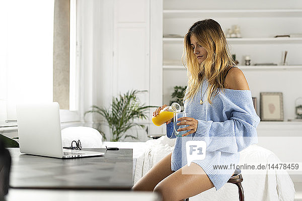 Junge Frau sitzt zu Hause im Schlafzimmer und gießt Orangensaft in ein Glas