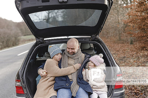 Lächelnder Vater mit zwei Kindern sitzt im Kofferraum am Straßenrand