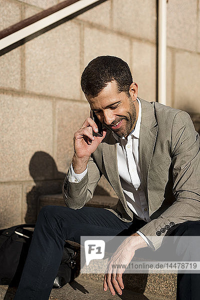 Lächelnder Geschäftsmann mit Tasche sitzt auf der Treppe und telefoniert mit dem Handy