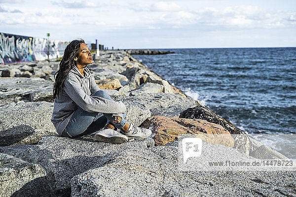 Junge Frau entspannt am Strand  auf Felsen sitzend  mit geschlossenen Augen
