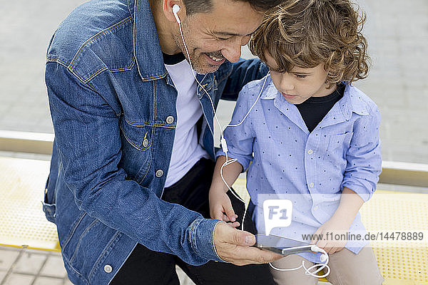Vater und Sohn sitzen an der Straßenbahnhaltestelle in der Stadt und teilen sich Handy und Ohrhörer