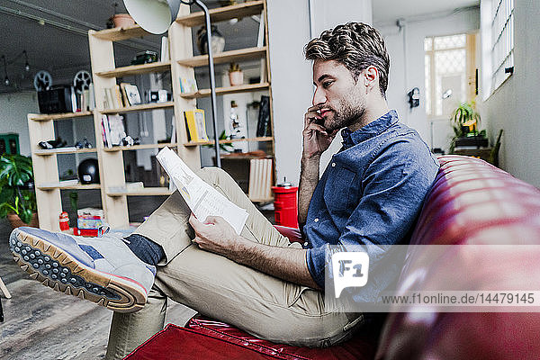 Businessman holding document talking on cell phone sitting on couch in loft office