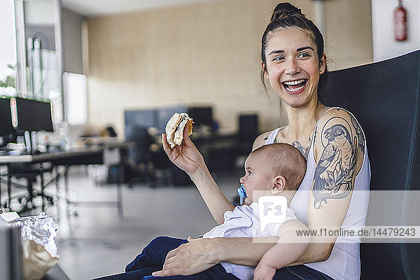 Young mother with tattoos sitting in office with her baby on lap  eating a sandwich