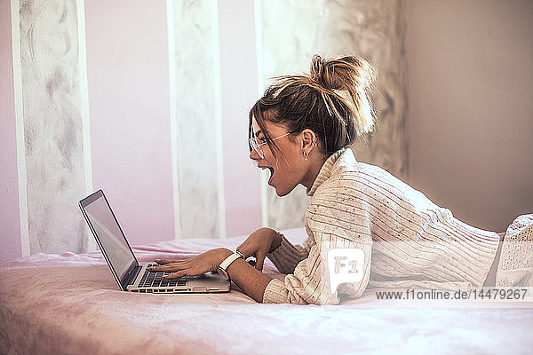 Überraschte junge Frau liegt mit Laptop auf dem Bett
