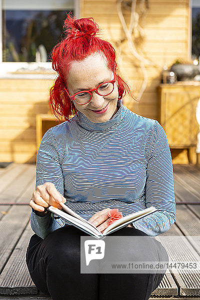 Porträt einer lächelnden älteren Frau mit rot gefärbten Haaren  die auf der Terrasse vor ihrem Haus sitzt und ein Buch liest
