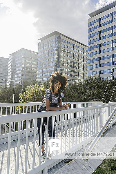 Junge Frau steht auf einer Brücke  benutzt ein Smartphone und hat Kopfhörer um den Hals