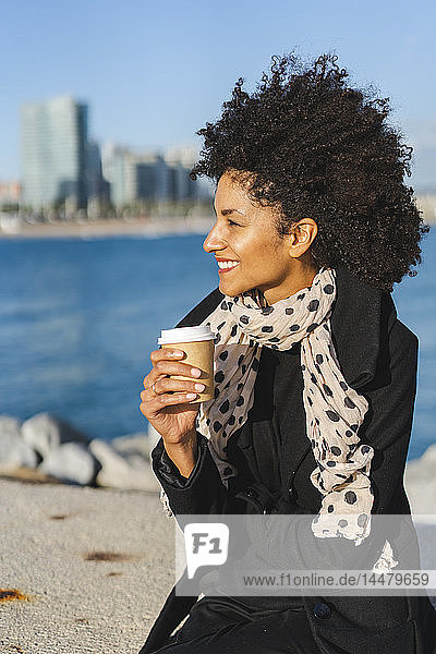 Spanien  Barcelona  lächelnde Frau sitzt auf einer Mauer mit Kaffee zum Mitnehmen