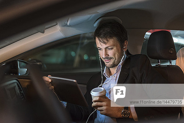 Businessman sitting in car at night  using digital tablet  drinking coffee