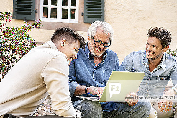 Three happy men of different age using laptop in garden