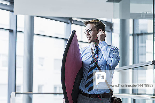Businessman with earphones carrying surfboard in office