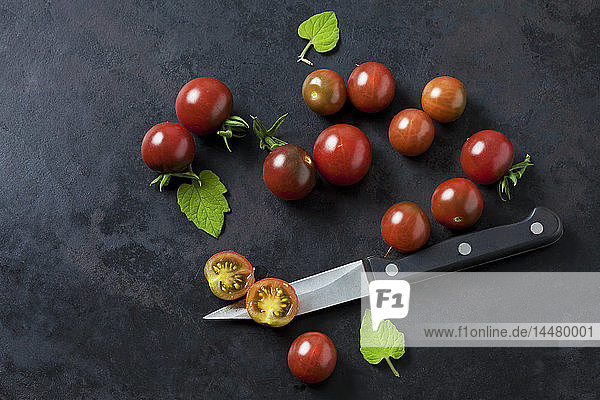 Ganze und in Scheiben geschnittene rispische Tomaten 'Black Cherry'  Blätter und Küchenmesser auf dunklem Grund