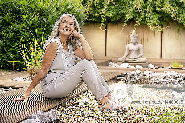 Lächelnde Frau mit langen grauen Haaren sitzt auf der Terrasse im Garten