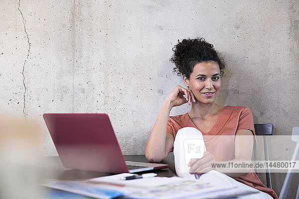 Porträt einer lächelnden Geschäftsfrau  die mit Dokumenten und Laptop am Tisch sitzt