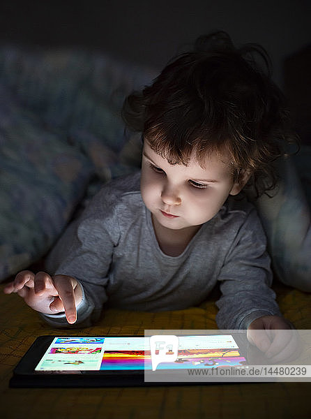 Kleines Mädchen benutzt digitales Tablett im Bett unter einer Decke