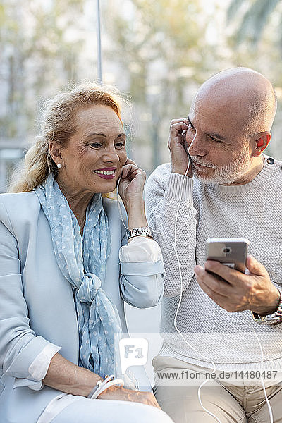 Spain  Barcelona  happy senior couple sharing smartphone with earbuds