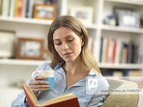 Junge Frau mit einem heißen Getränk  die sich zu Hause beim Lesen eines Buches entspannt