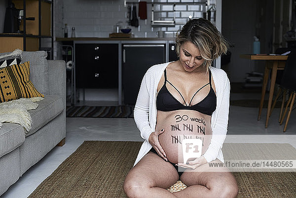Schwangere Frau sitzt zu Hause auf dem Boden und schaut auf Strichmarken auf ihrem Bauch