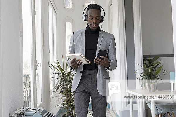 Porträt eines jungen Geschäftsmannes mit Kopfhörern und Notizbuch im Büro beim Blick auf das Handy