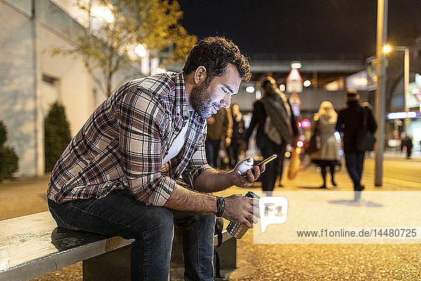 Großbritannien  London  lächelnder Mann sitzt nachts auf einer Bank und schaut auf sein Telefon