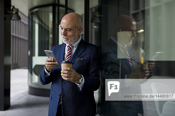 Leitender Geschäftsmann mit Handy und Kaffee zum Mitnehmen an der Glasfront