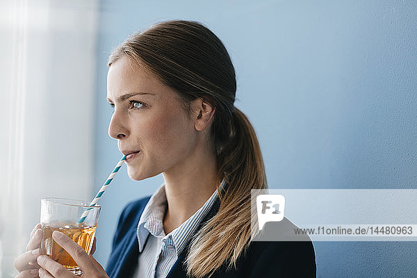 Porträt einer jungen Geschäftsfrau vor blauem Hintergrund  die mit einem Strohhalm Eistee trinkt