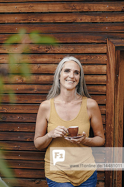 Porträt einer glücklichen reifen Frau mit Kaffeetasse vor einer Holzfassade
