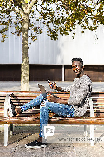 Young man sitting on a bench in the city  using laptop and smartphone