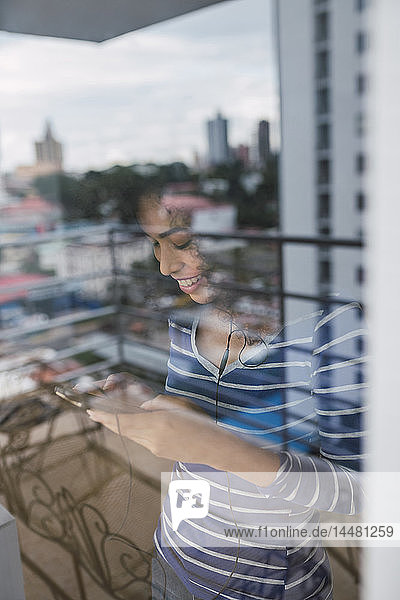 Lächelnde junge Frau hinter Fensterscheibe mit Smartphone und Kopfhörern