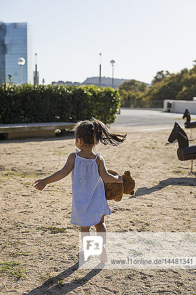 Rückansicht eines kleinen Mädchens mit Teddy auf einem Spielplatz