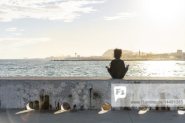 Spanien  Barcelona  Rückenansicht einer Frau  die an einer Wand vor dem Meer sitzt und meditiert
