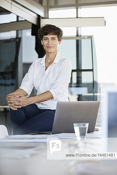 Portrait of confident businesswoman sitting on desk in office with laptop