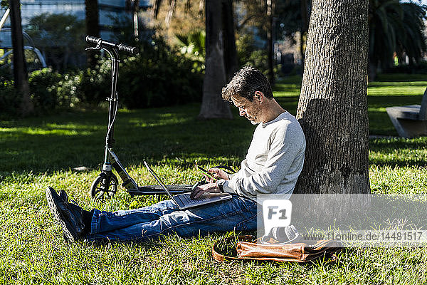 zufälliger Geschäftsmann  der auf einem Rasen in einem Park sitzt und einen Laptop benutzt