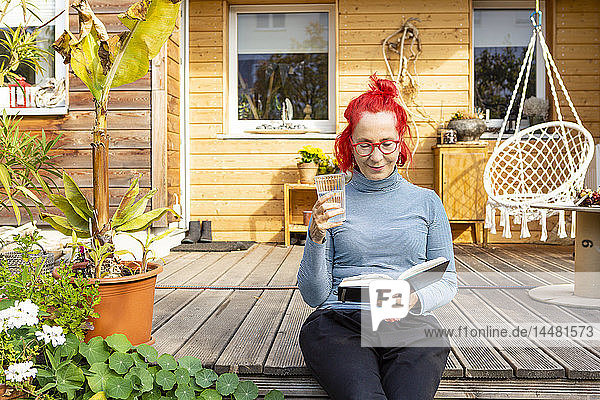 Porträt einer lächelnden älteren Frau mit rot gefärbten Haaren  die auf der Terrasse vor ihrem Haus sitzt und ein Buch liest