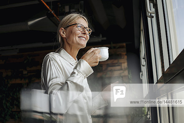 Lächelnde Geschäftsfrau mit einer Tasse Kaffee  die aus dem Fenster schaut