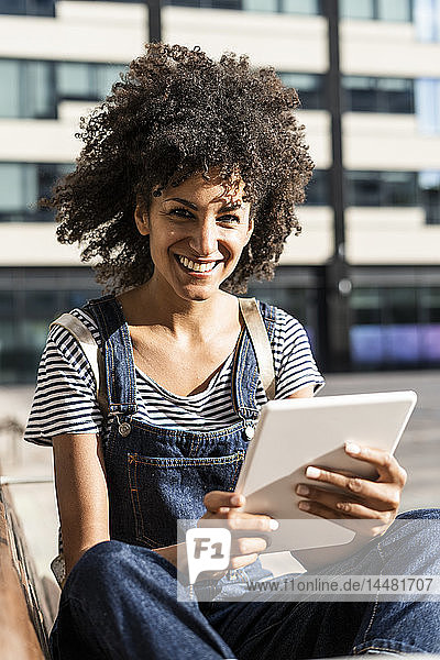 Mittlere erwachsene Frau mit lockigem Haar  die auf einer Bank sitzt und ein digitales Tablett benutzt