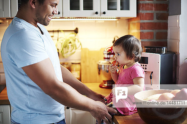 Smiling father looking at baby girl eating a tangerine on counter in kitchen at home