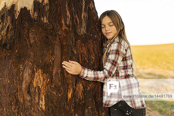 Porträt eines zufriedenen blonden Mädchens  das einen Baumstamm umarmt