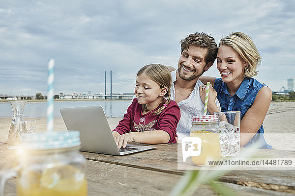 Deutschland  Düsseldorf  glückliche Familie mit Tochter mit Laptop auf Holztisch am Rheinufer