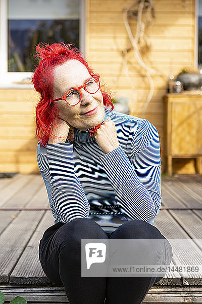 Porträt einer lächelnden älteren Frau mit rot gefärbten Haaren  die auf der Terrasse vor ihrem Haus sitzt und sich entspannt