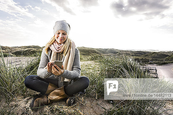 Lächelnde Frau sitzt in den Dünen und benutzt Handys