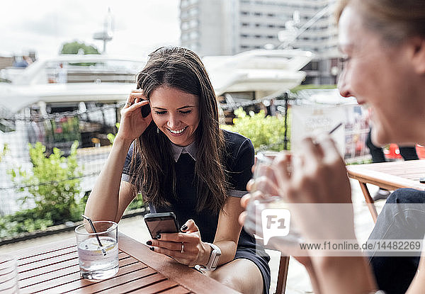 Zwei glückliche Frauen mit Smartphone und Getränken auf einer Terrasse