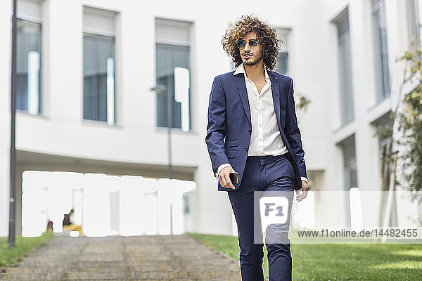Portrait of young fashionable businessman with curly hair wearing blue suit and sunglasses