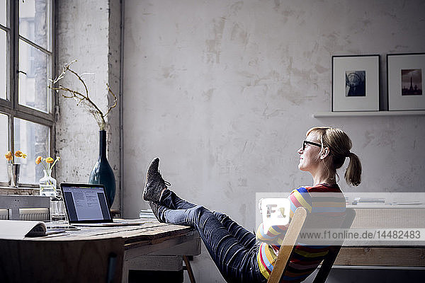Smiling woman sitting with feet up at desk in a loft looking through window