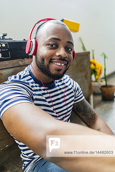 Porträt eines lächelnden jungen Mannes mit Kopfhörern  der in einem Loft auf dem Boden sitzt