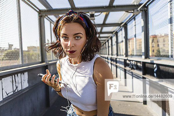 Porträt einer jungen Frau mit Ohrstöpseln und Handy auf einer Brücke