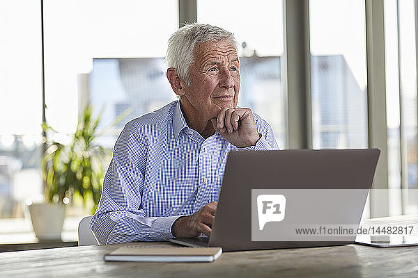 Porträt eines nachdenklichen älteren Mannes  der mit Laptop am Tisch sitzt und in die Ferne schaut