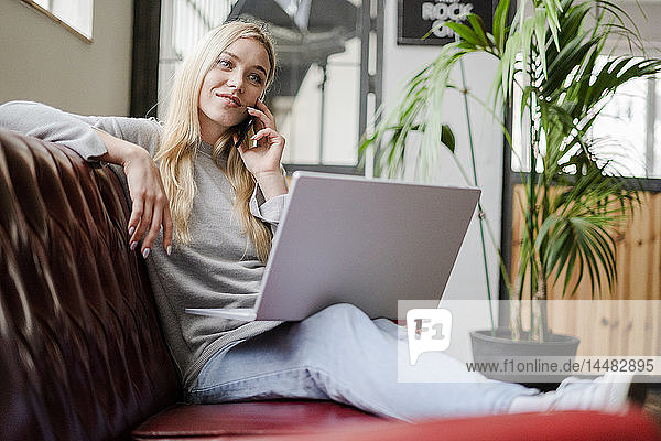 Lächelnde junge Frau sitzt auf der Couch mit Laptop und telefoniert mit dem Handy