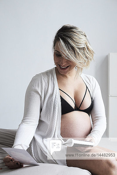 Lächelnde Schwangere  die zu Hause auf dem Bett sitzend Ultraschallbilder betrachtet