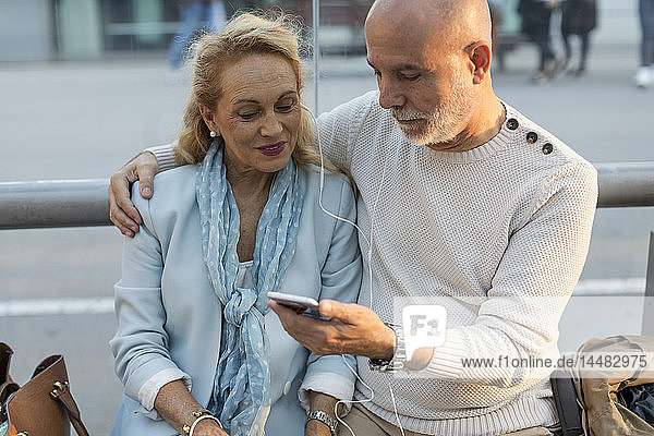 Spain  Barcelona  senior couple sitting at tram stop in the city sharing smartphone with earbuds